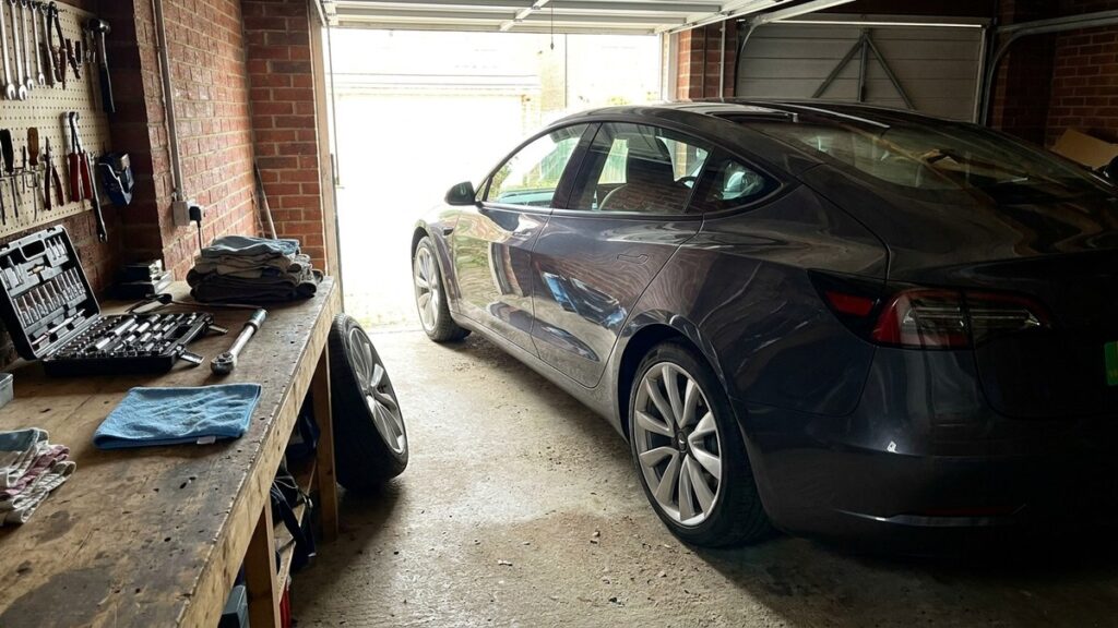 A modern electric car parked in a domestic garage beside a workbench laid out with a socket set, torque wrench and cloths
