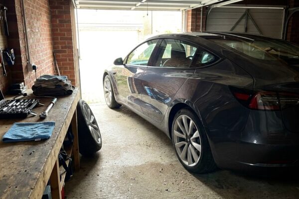 A modern electric car parked in a domestic garage beside a workbench laid out with a socket set, torque wrench and cloths