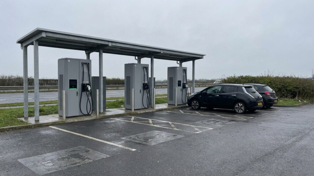 A roadside rapid-charging hub under a steel canopy on an overcast day, two cars charging at the far bays