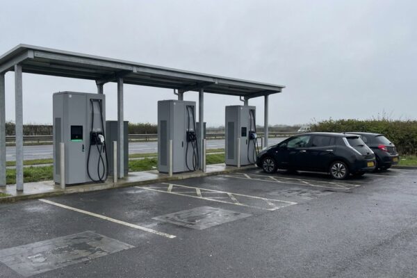 A roadside rapid-charging hub under a steel canopy on an overcast day, two cars charging at the far bays