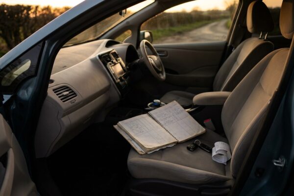 A ruled paper mileage log lies open on the passenger seat of a parked electric car at dusk, beside car keys and a folded receipt
