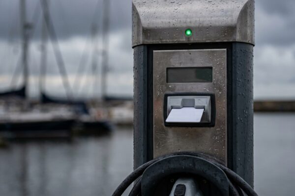 Rain beads on a quayside EV charge post, a prepay card seated in its reader and a green indicator lit, moored yachts blurred behind